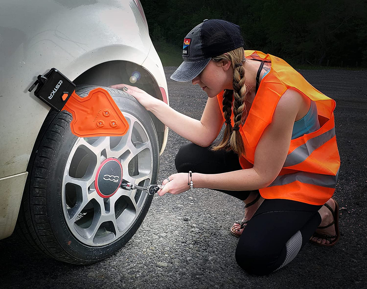  A woman in an orange safety vest uses a portable tool to change a car tire on the roadside.